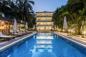 A swimming pool with lounge chairs and umbrellas, surrounded by palm trees and a multi-story building with balconies. - Safira River Front Resort (Morjim)