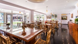 A spacious dining area with glass tables and wicker chairs, large windows offering views of a garden, and a modern ceiling design. - Safira River Front Resort (Morjim)