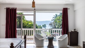 A bedroom with a large bed, two white chairs, a small table, and a view of the sea through a sliding glass door. - Safira River Front Resort (Morjim)