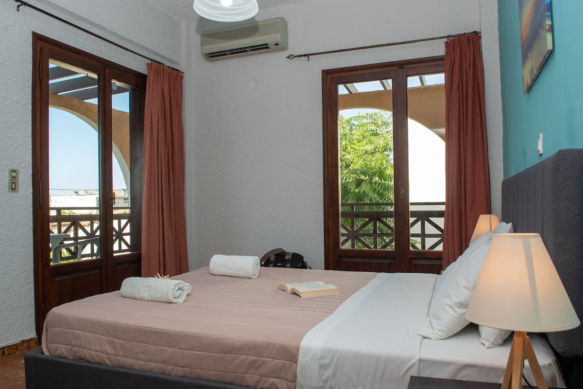 A hotel room with a bed, a lamp, and a view of greenery through the windows.