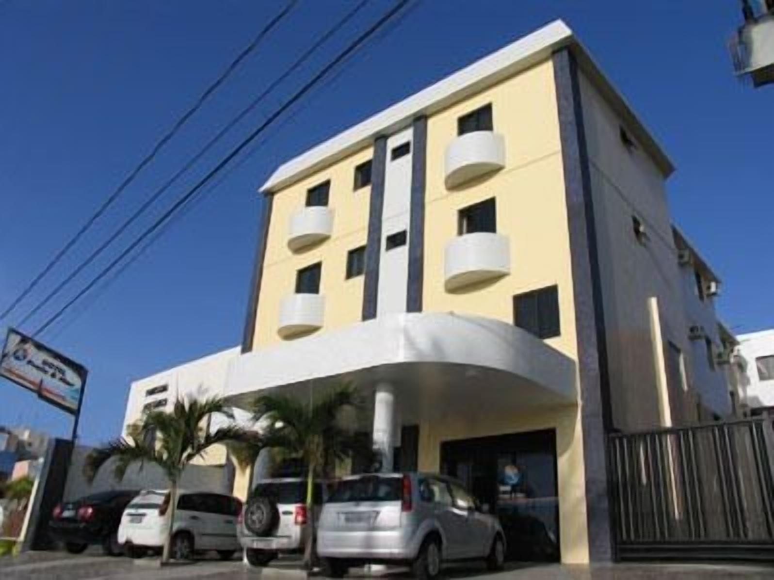 A multi-story hotel with a flat facade, balconies, and a parking area with several cars.