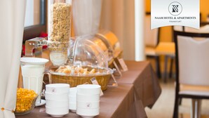 A buffet setup with various food items and containers, including a cereal dispenser and a basket of bread. - Naam Hotel & Apartment Frankfurt City-messe Airpor (Frankfurt)