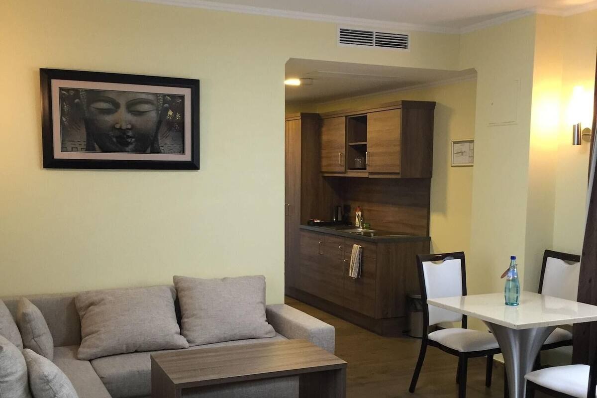 Sala de estar com sofá, mesa de centro e mesa de jantar com cadeiras. Há também uma área de cozinha com armários e pia.