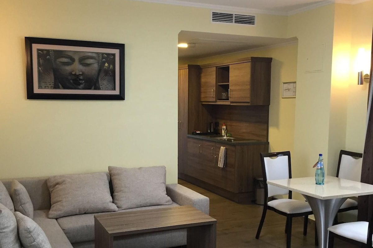A living room with a sofa, a coffee table, and a dining table with chairs. There is a kitchen area with cabinets and a sink.