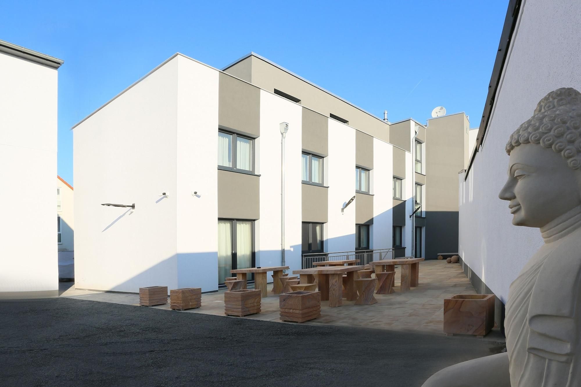 A modern building with a flat facade, multiple windows, and a statue of a Buddha. There are wooden benches and tables arranged outside.