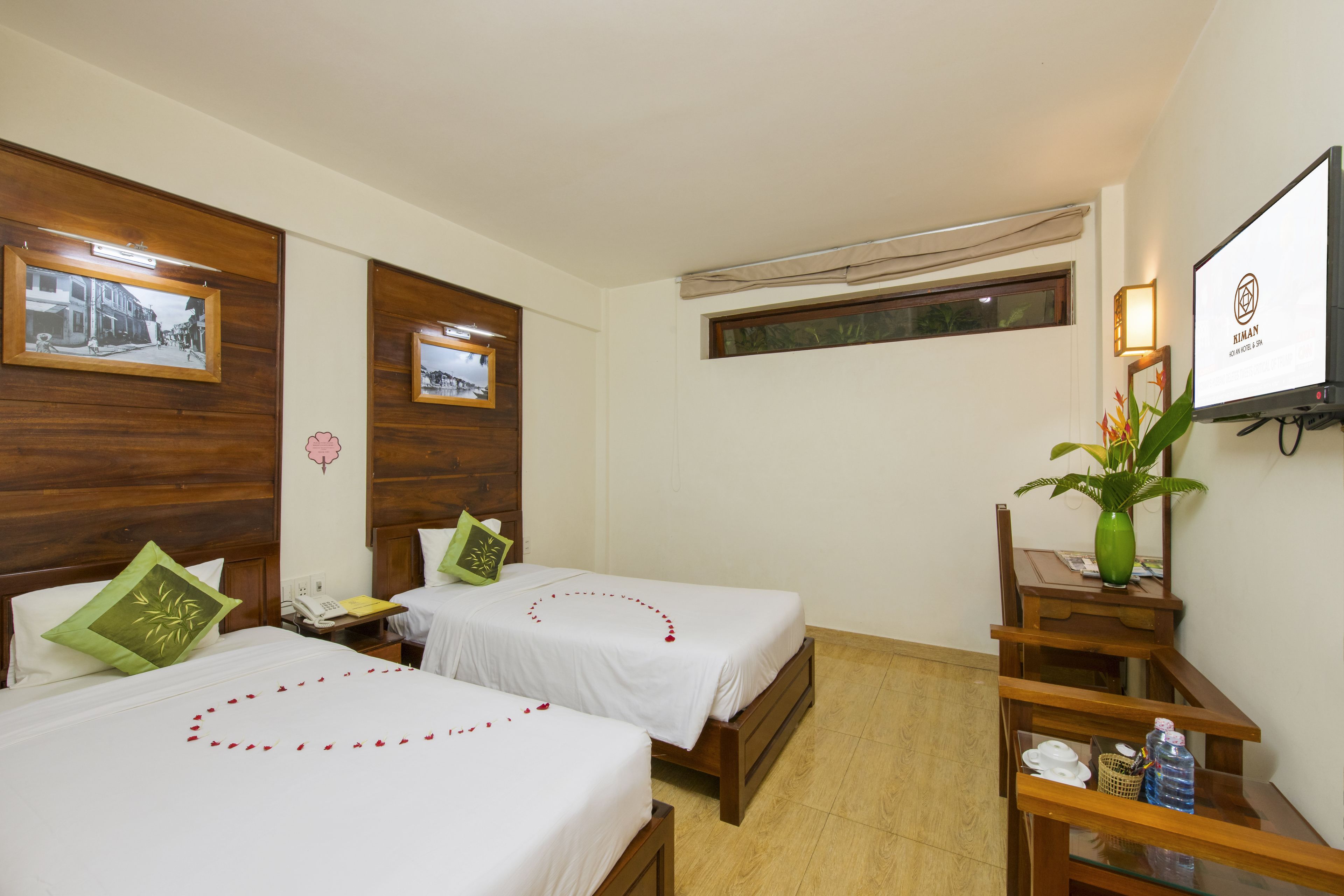 A hotel room with two beds, wooden headboards, a small desk with a lamp, a TV, and a window with a view of the street.