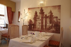 Un comedor con una mesa dispuesta para dos personas, un cuadro histórico grande en la pared y una ventana con cortinas.