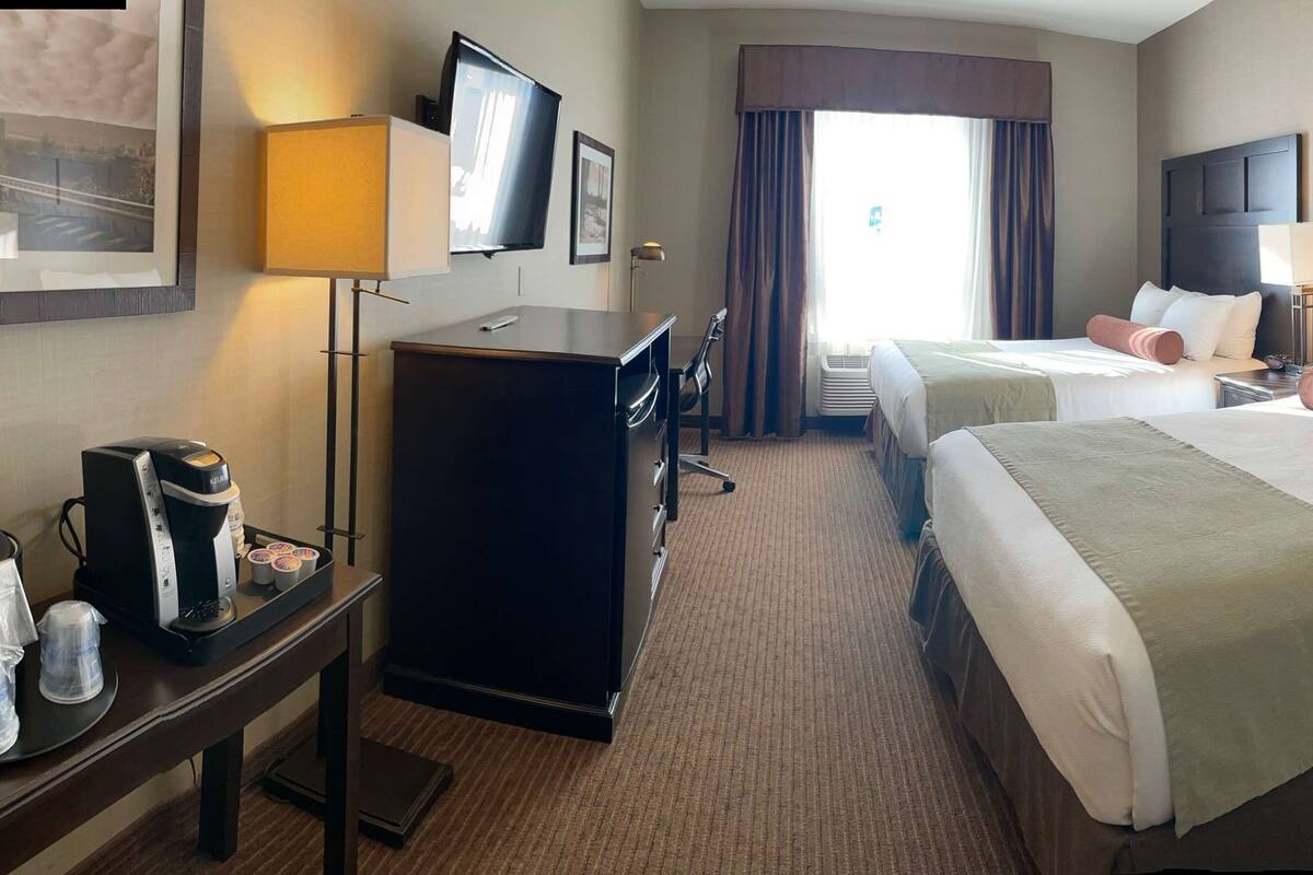 A hotel room with two beds, a desk with a coffee maker, a TV, and a large window with curtains.