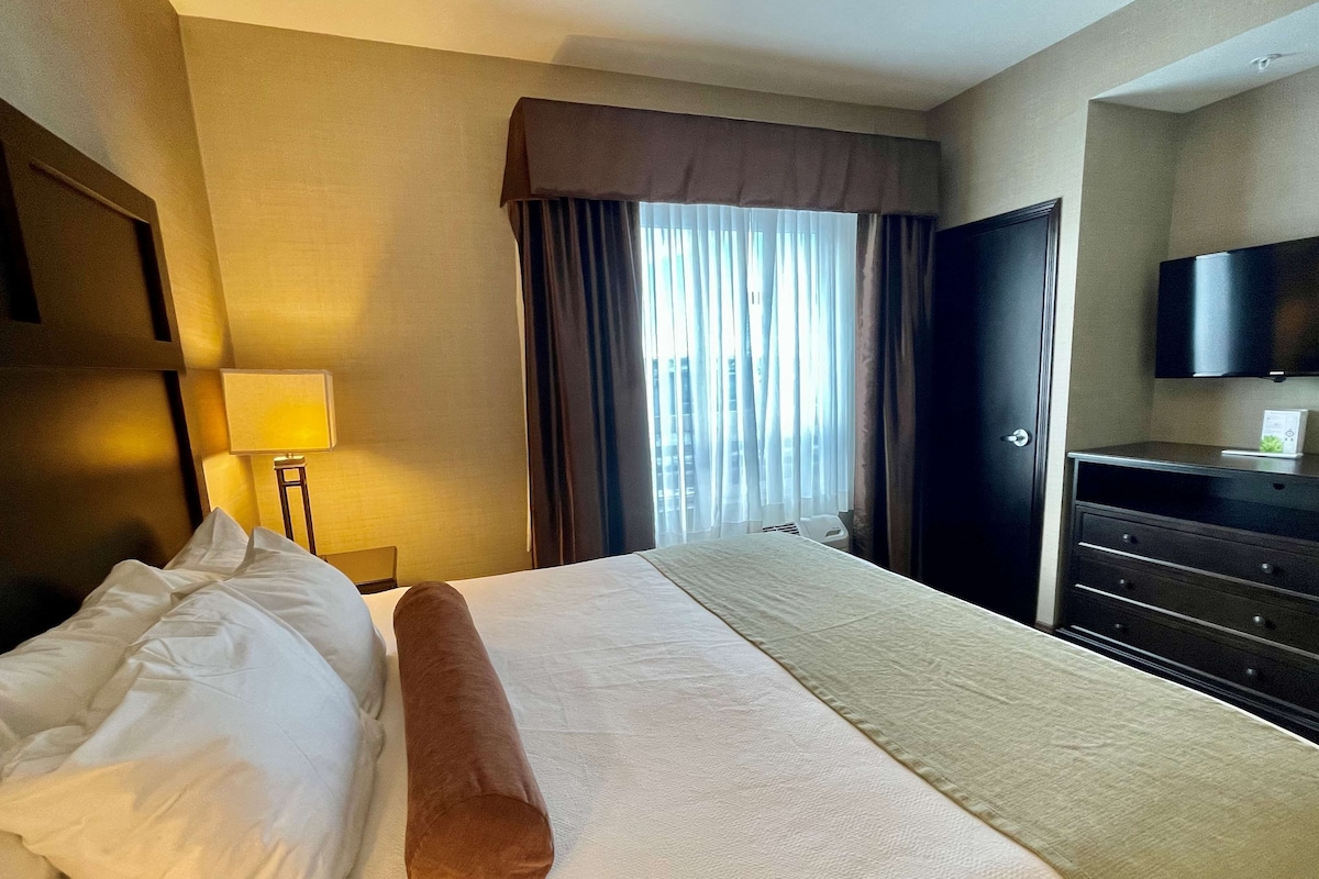 A hotel room with a bed, a nightstand, a lamp, a TV, and a window with curtains.
