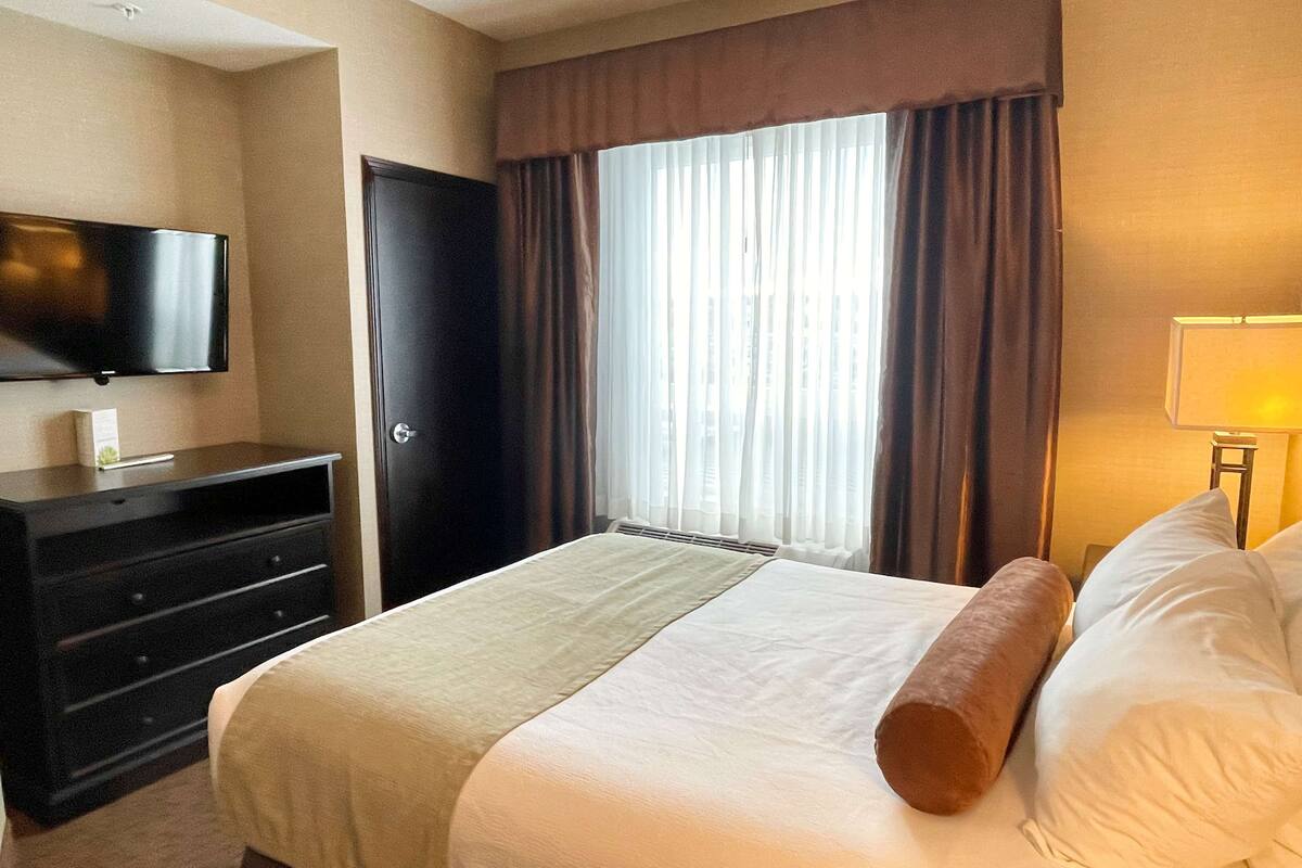 A hotel room with a bed, a TV mounted on the wall, a dresser, and a window with curtains.