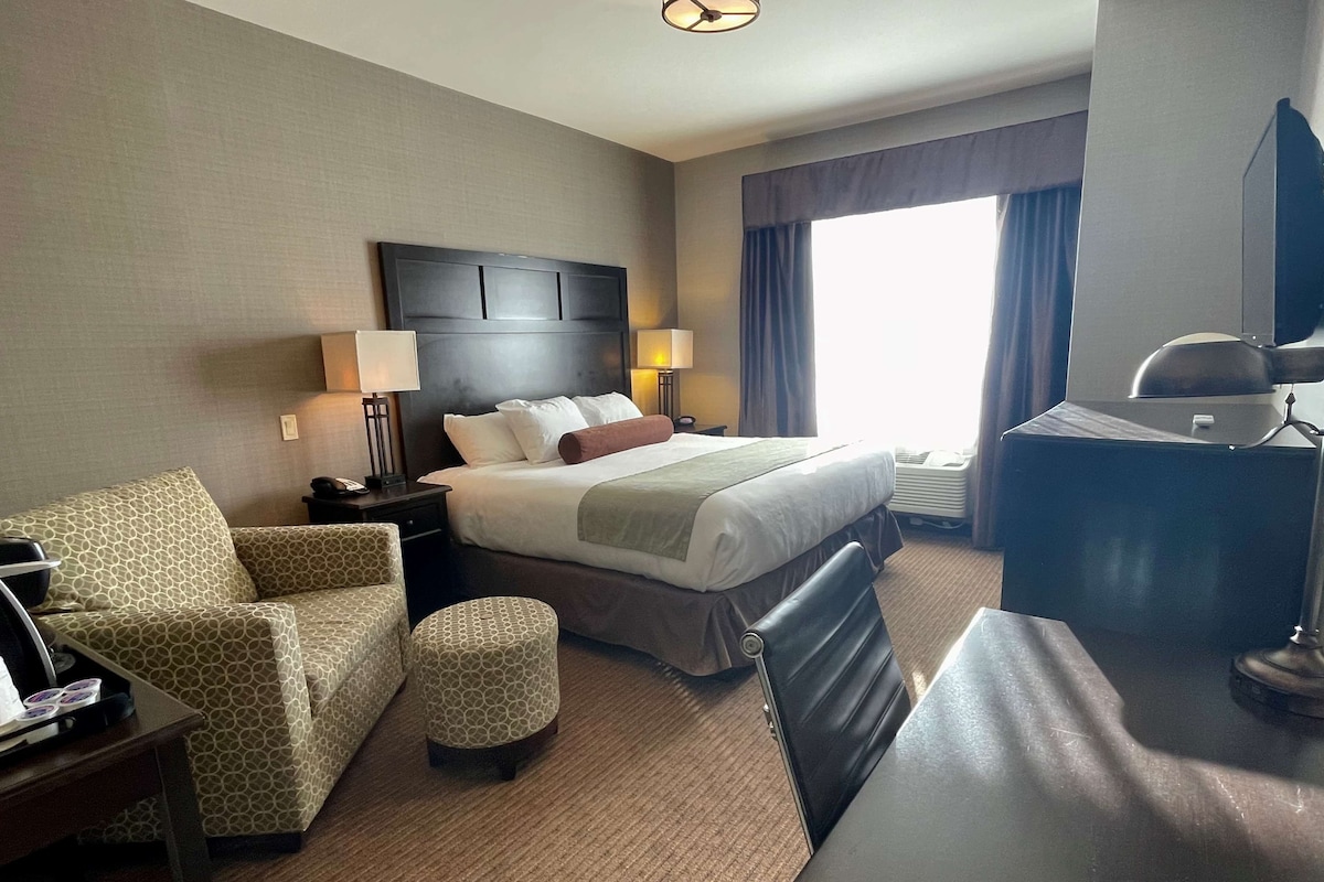 A hotel room with a bed, a desk, a chair, a small ottoman, and a desk lamp.