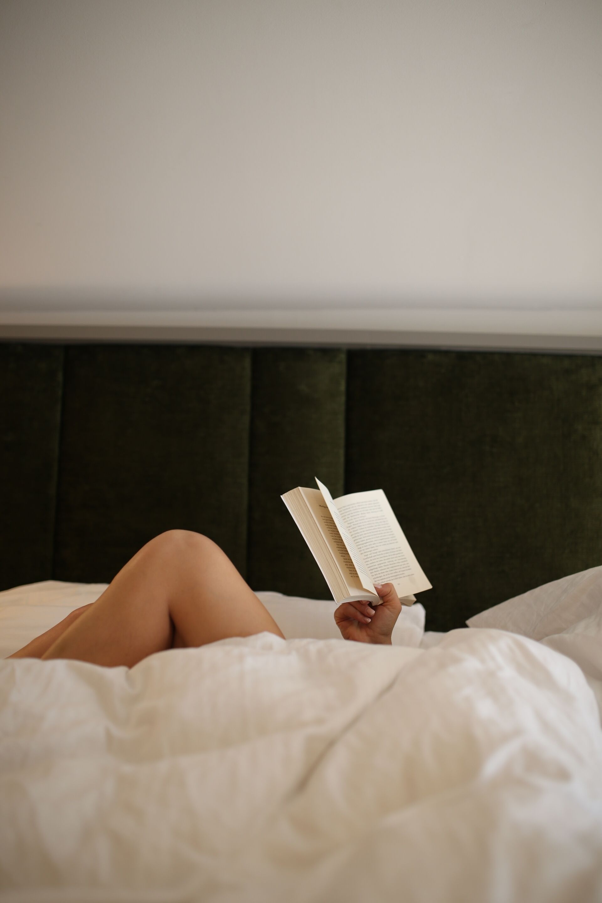 Una persona recostada en una cama con un libro, en una habitación con un cabecero oscuro y ropa de cama blanca.