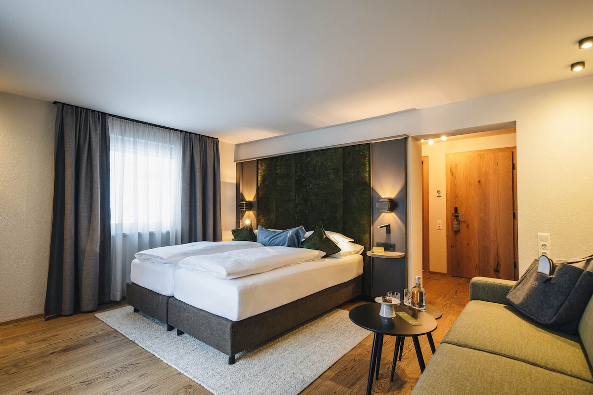 A modern hotel room with a large bed, a small sofa, a round table, and a wooden door.
