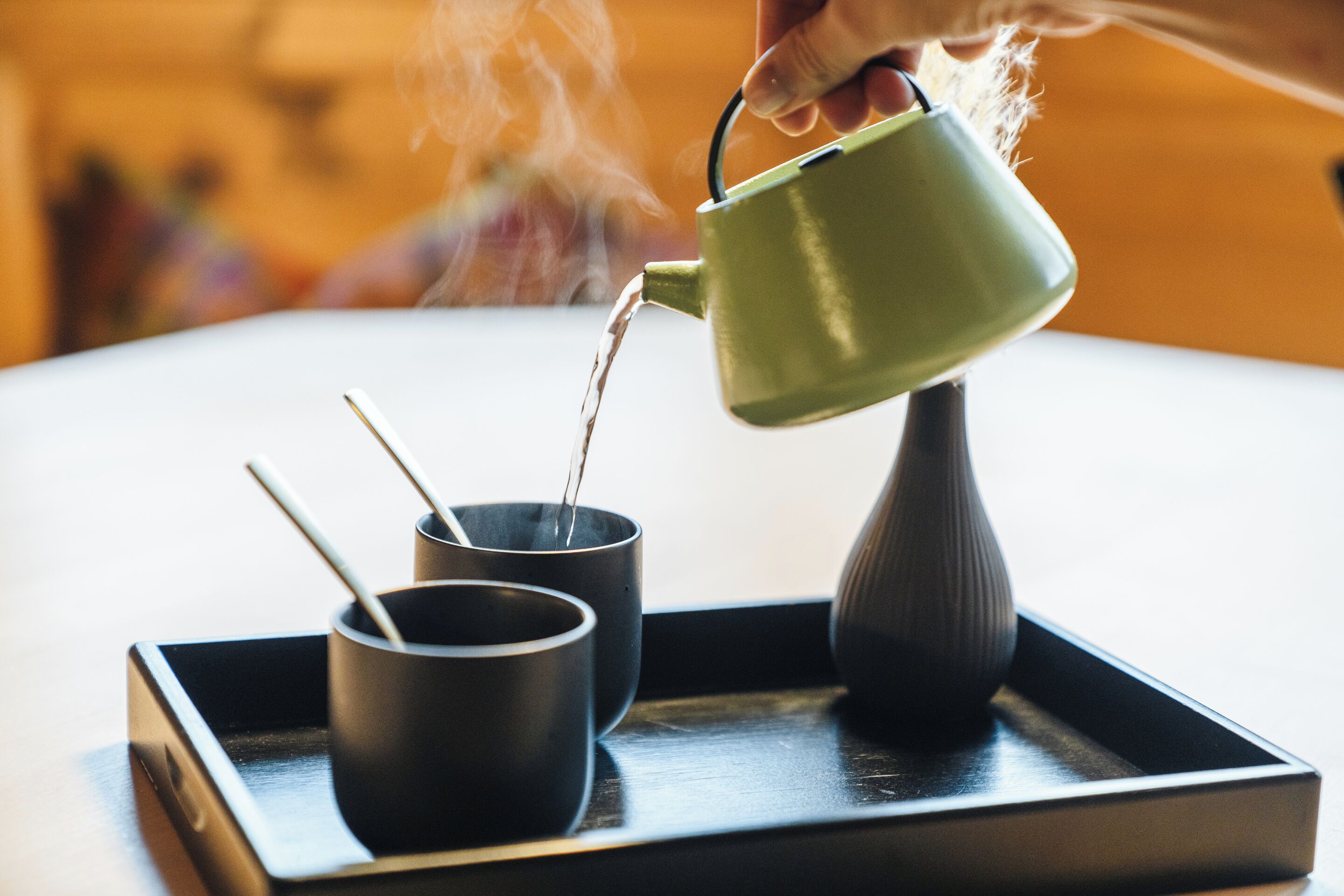 Una mà abocant aigua d'una gerra verda en dues tasses negres sobre un plat.