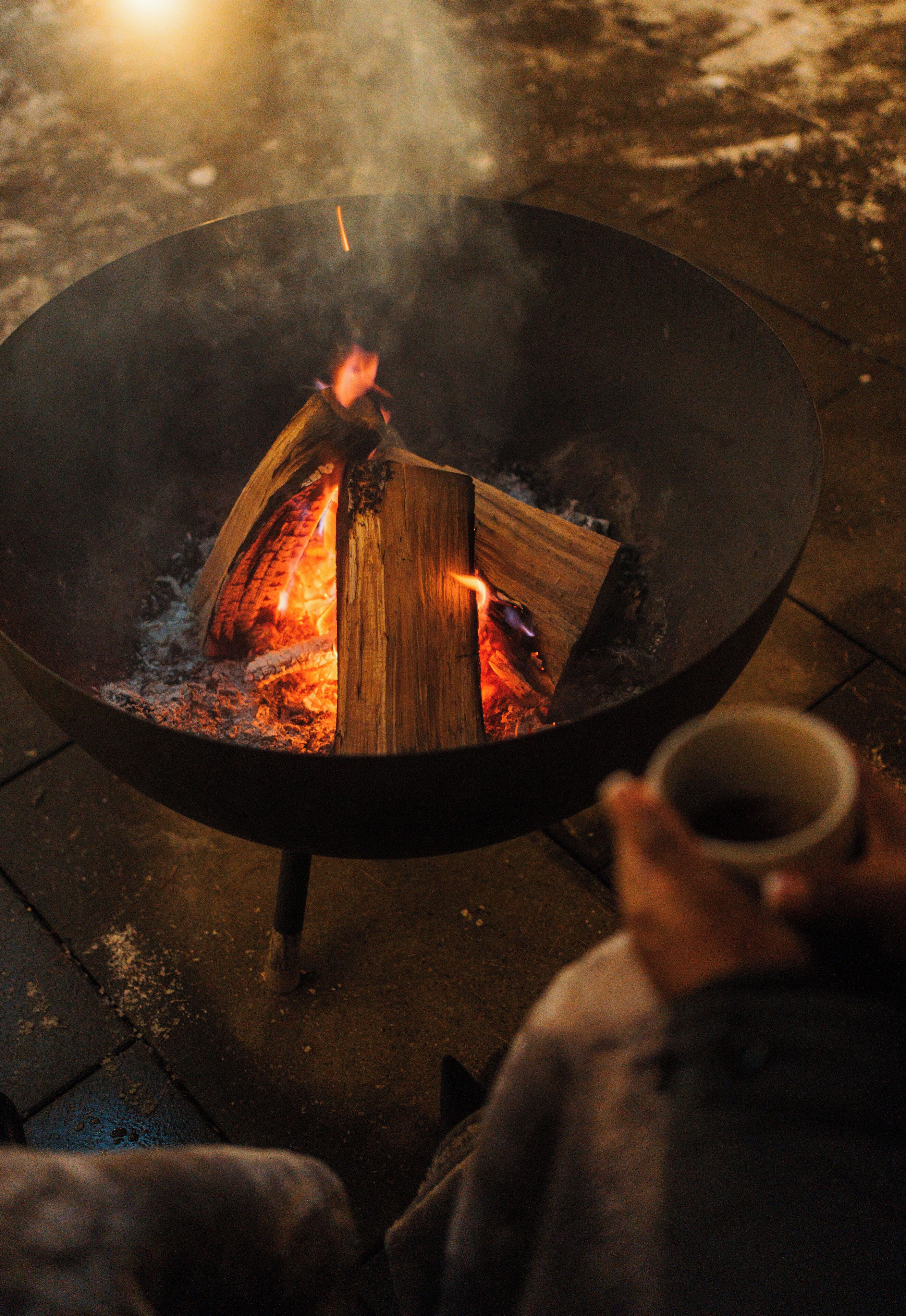 A circular outdoor fire pit with logs and flames, a person holding a mug, and a paved ground.