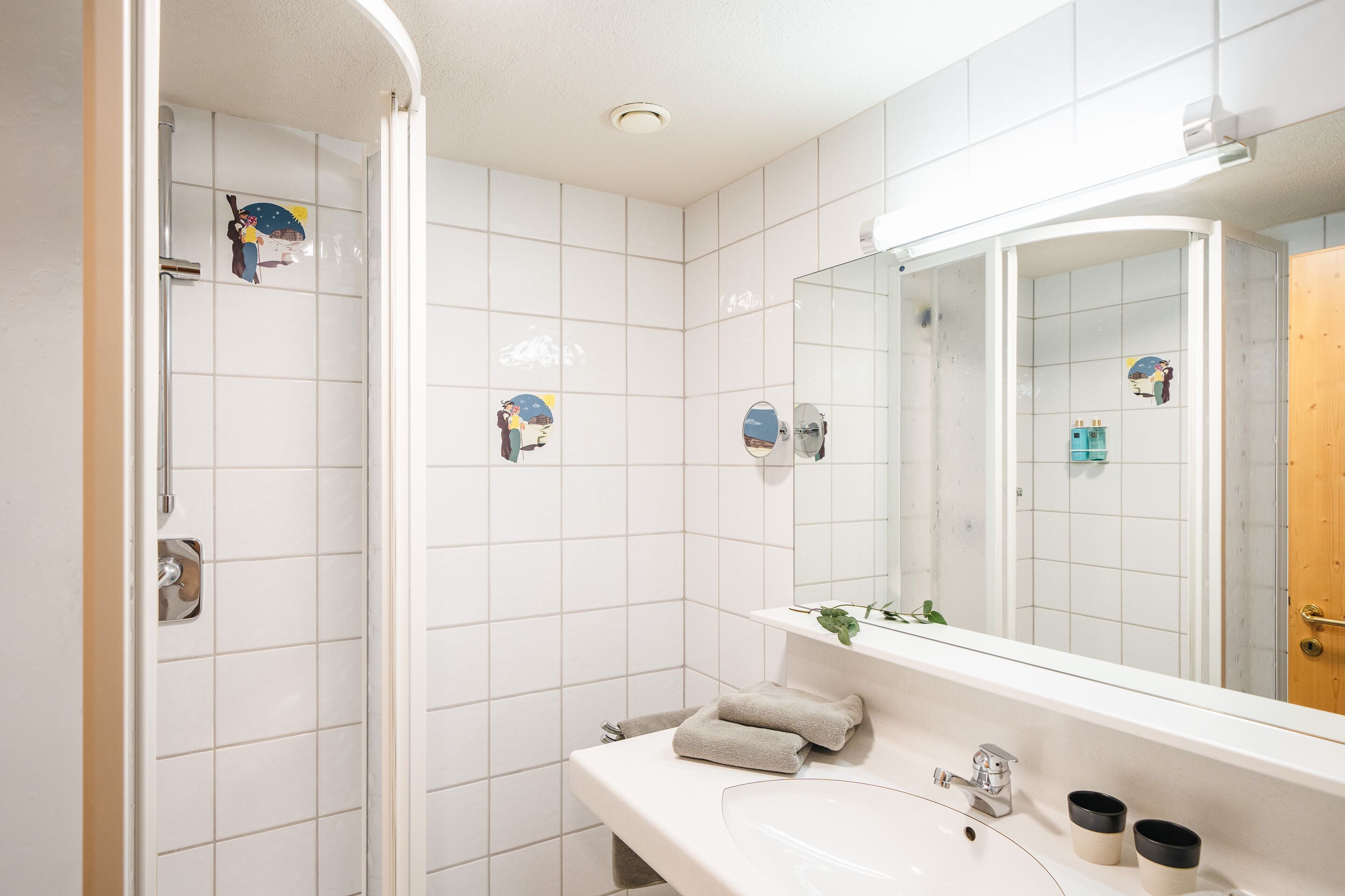A bathroom with white tiled walls, a sink, a mirror, and a shower.