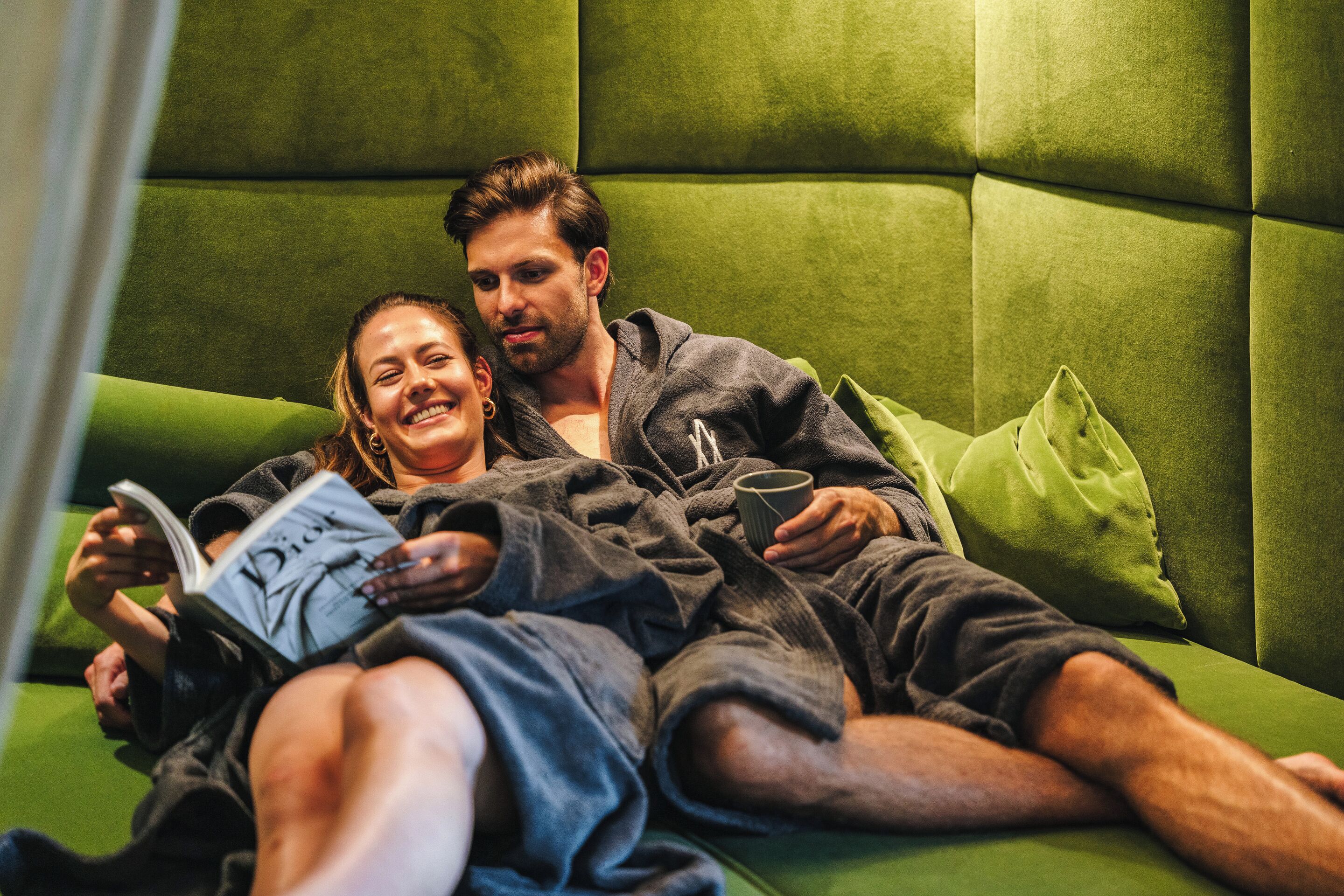 A couple lying on a green upholstered sofa with pillows, reading a book.