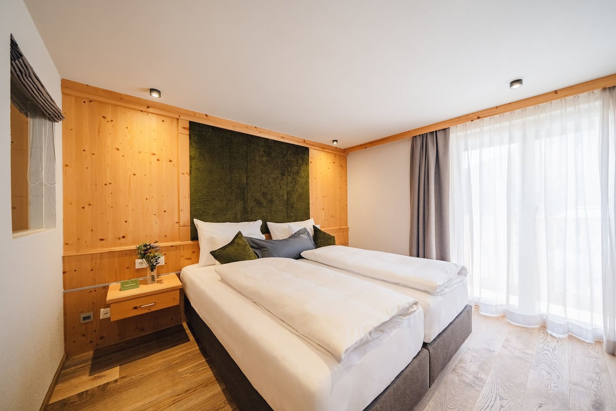 A double bed with white bedding, a wooden headboard, a nightstand with a lamp, and a window with sheer curtains.