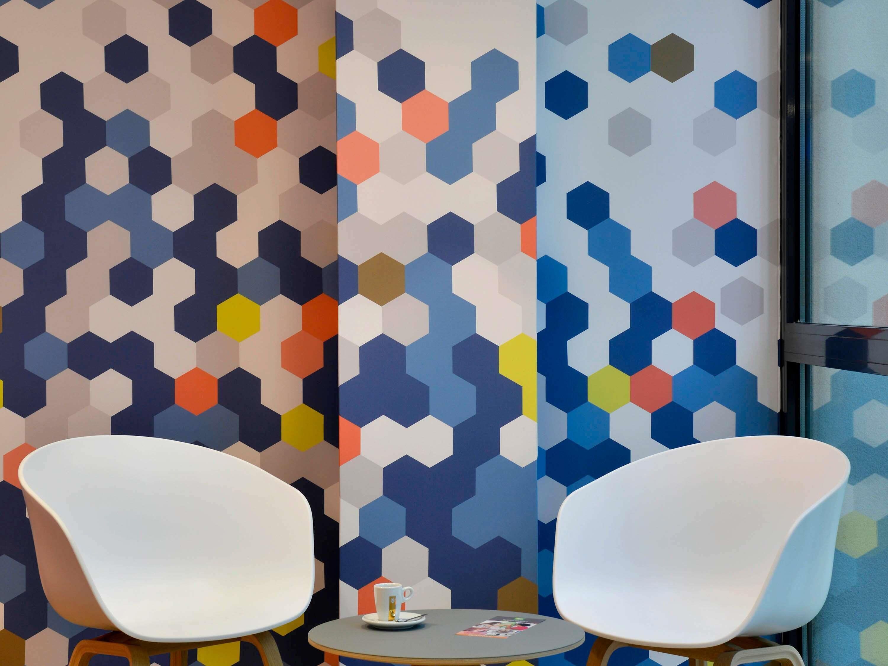 Una habitación con dos sillas blancas, una pequeña mesa redonda y un pared con un patrón hexagonal de colores.