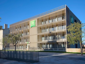 A modern hotel building with a vertical facade, multiple balconies, and a green sign indicating it is an ibis hotel.