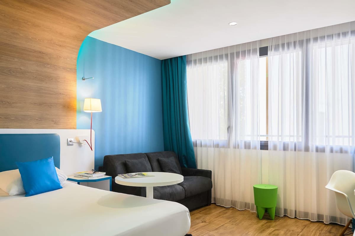 A hotel room with a bed, a small table, a sofa, a green stool, and a window with sheer curtains.