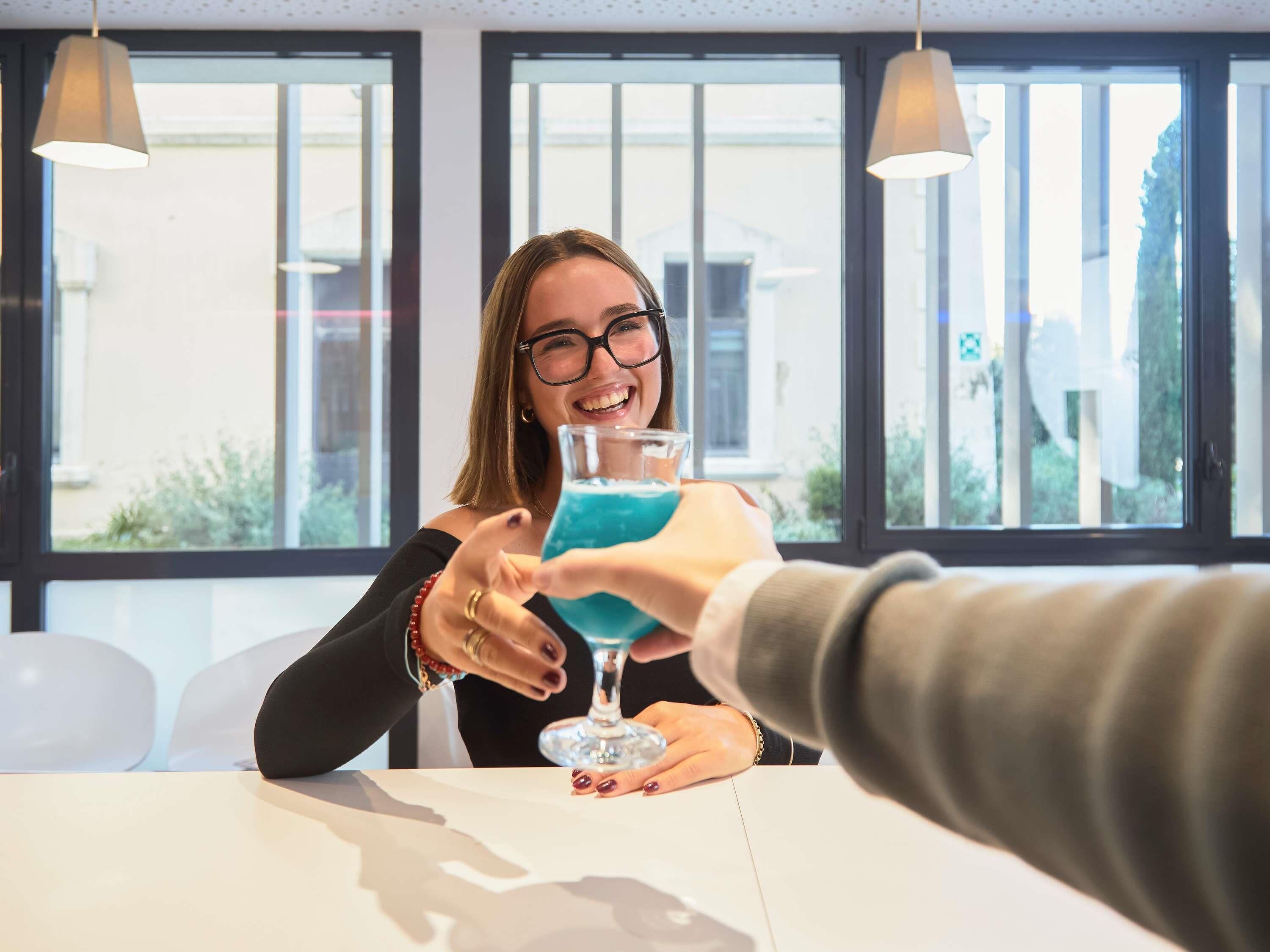 A person holding a glass with a blue drink, seated at a table with another person extending a hand towards them.