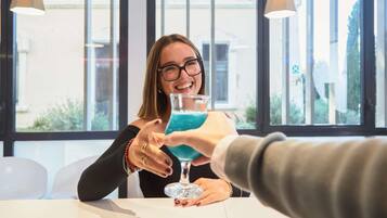 A person holding a glass with a blue drink, seated at a table with another person extending a hand towards them.