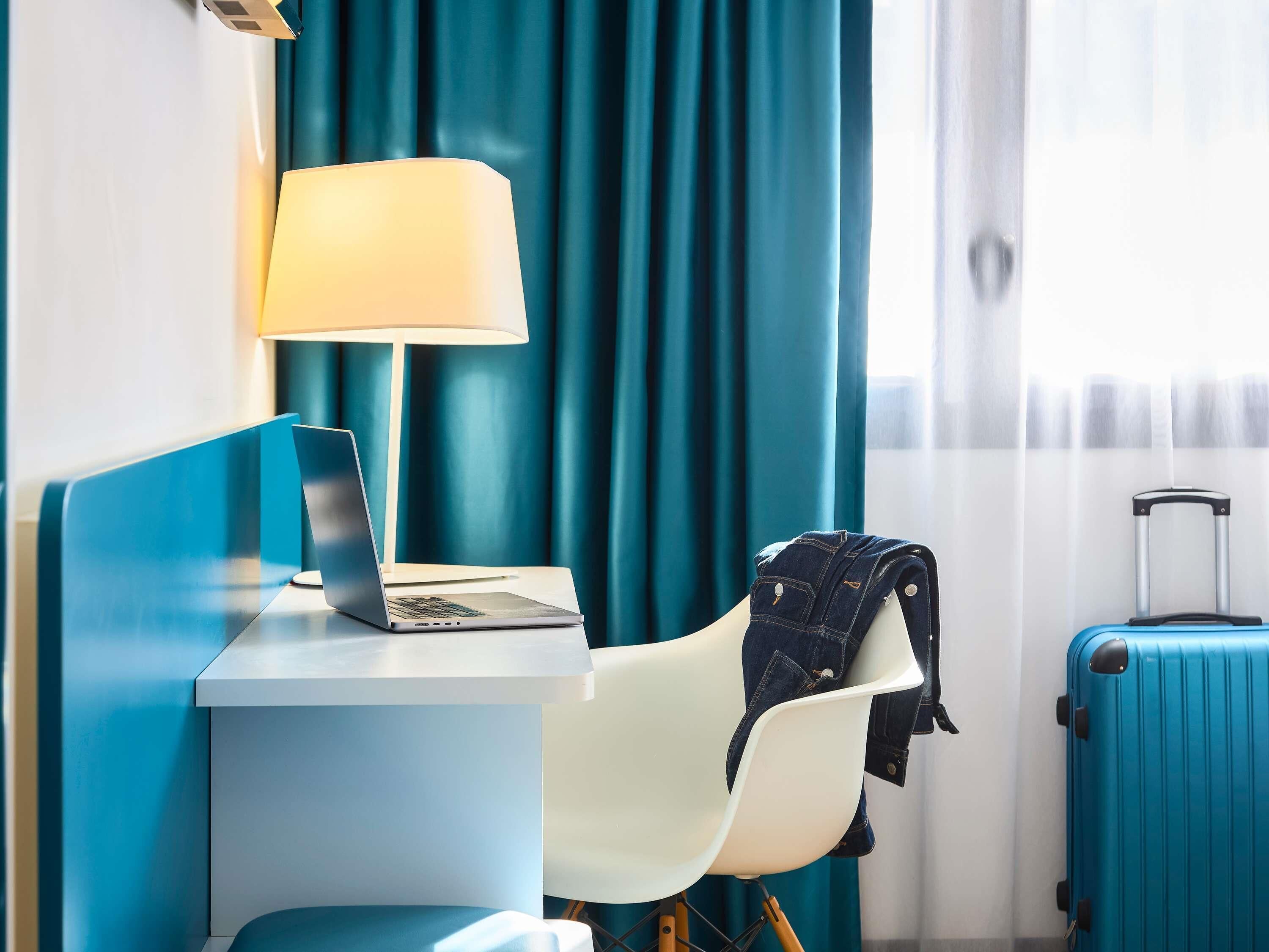 A hotel room with a desk, a laptop, a chair, a lamp, a jacket on the chair, and a suitcase.