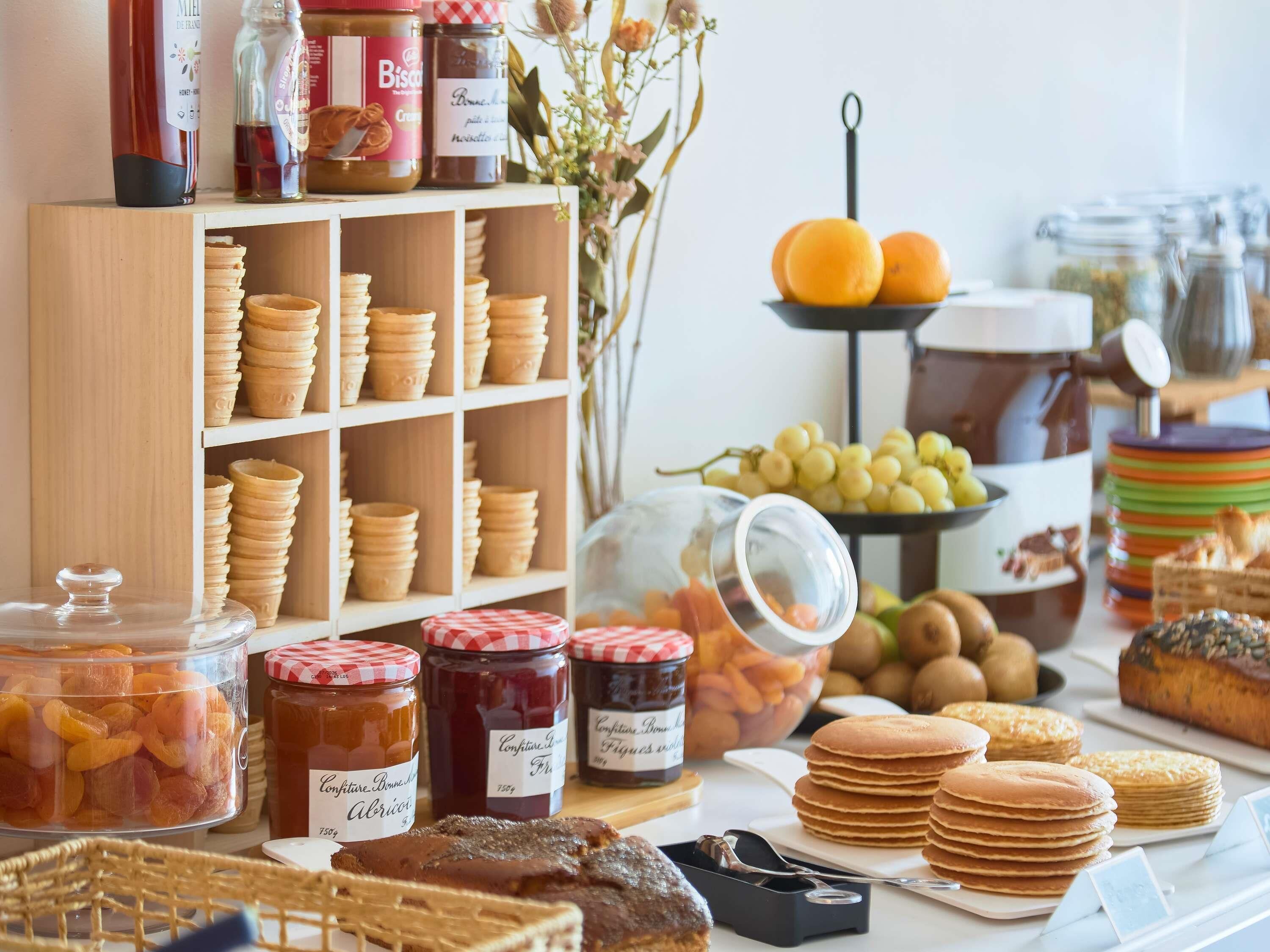 Eine Auswahl an Speisen und Beilagen präsentiert sich, darunter Gläser mit Marmelade, eine Schale mit Trauben und stapelten Pfannkuchen.