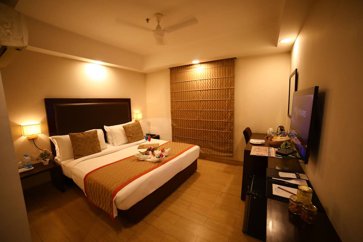 A hotel room with a bed, bedside table, lamp, TV, and a desk with a chair.