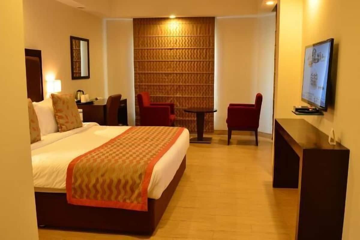 A hotel room with a bed, a desk, a chair, a TV, and a wall-mounted shelf.