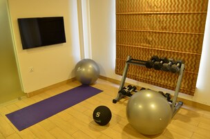 A room with a flat-screen TV mounted on the wall, a purple exercise mat, two large exercise balls, and a dumbbell rack with weights.