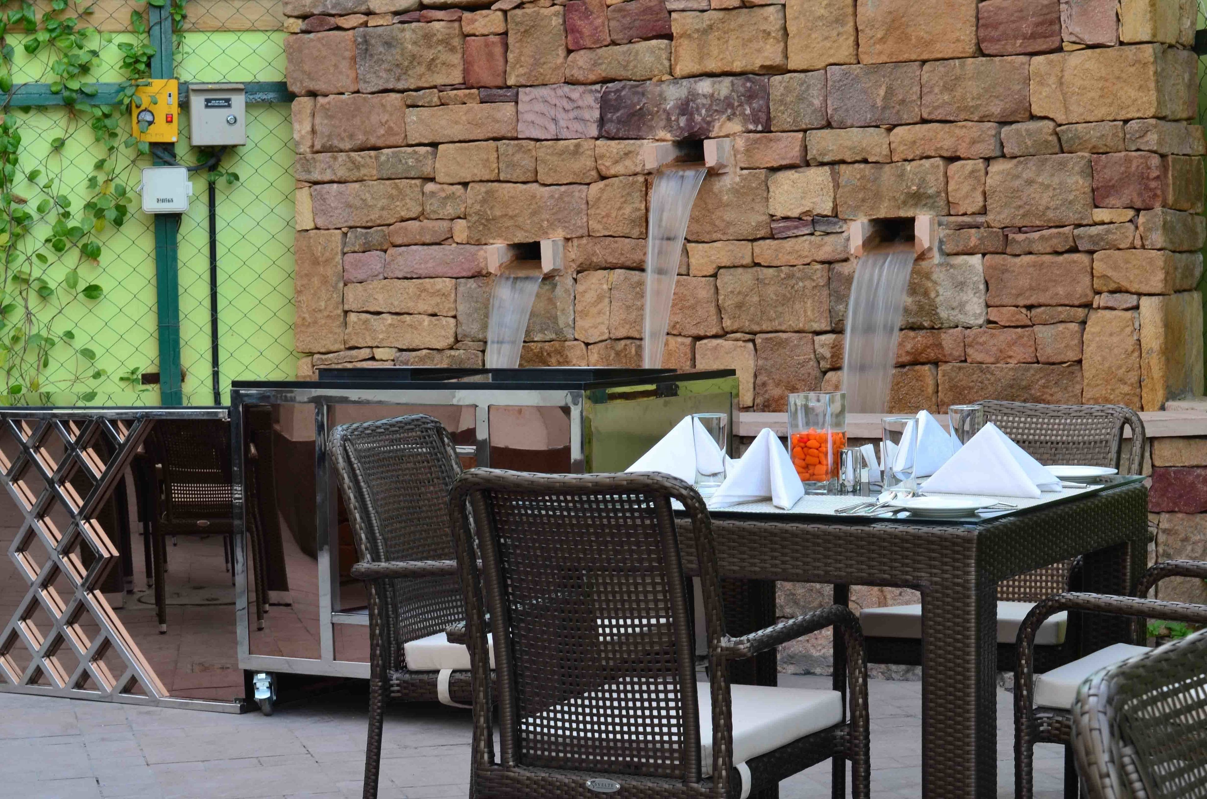 An outdoor dining area with wicker chairs and a table set for two, a stone wall with a water feature, and electrical boxes on the wall.