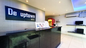 Ein Hotelrezeptionstresen mit einem Schild, das "De Uptown Hotel" zeigt, ein Coca-Cola-Kühlschrank und ein Flachbildfernseher auf einem Regal.
