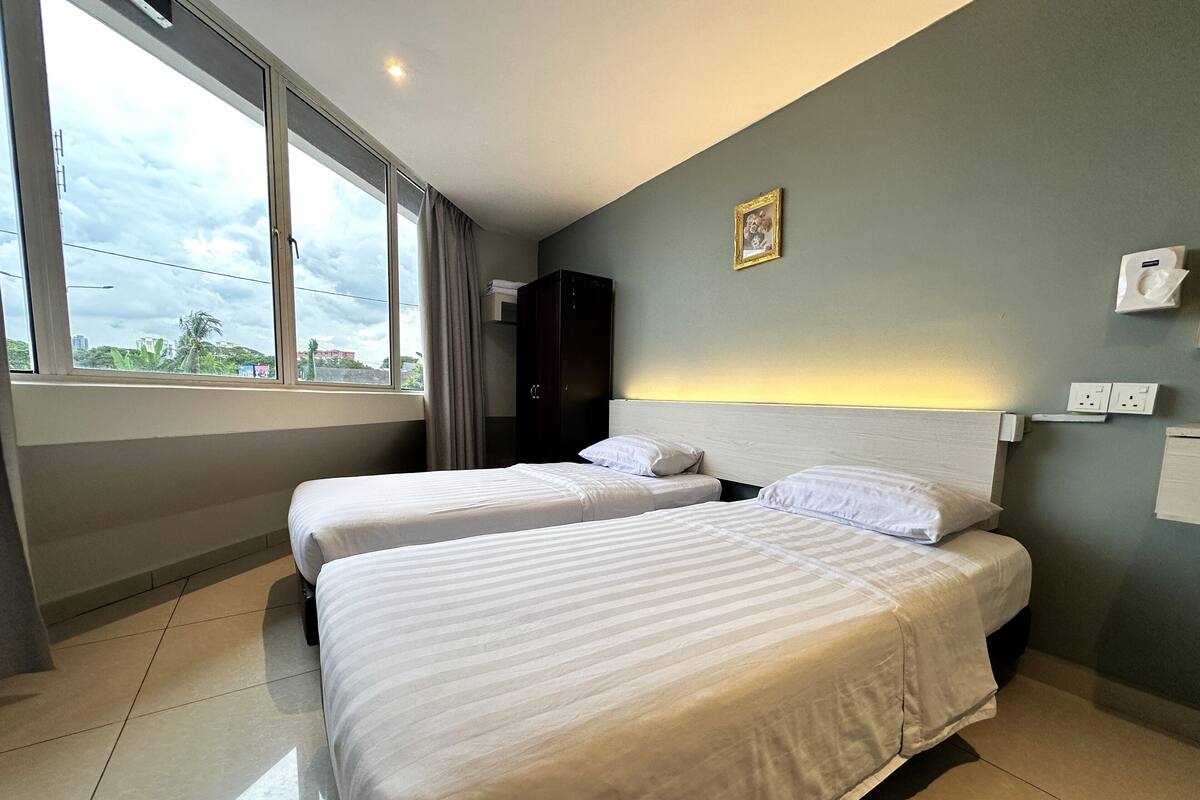 Une chambre d’hôtel avec deux lits, une grande fenêtre, une armoire sombre et un luminaire fixé au mur.