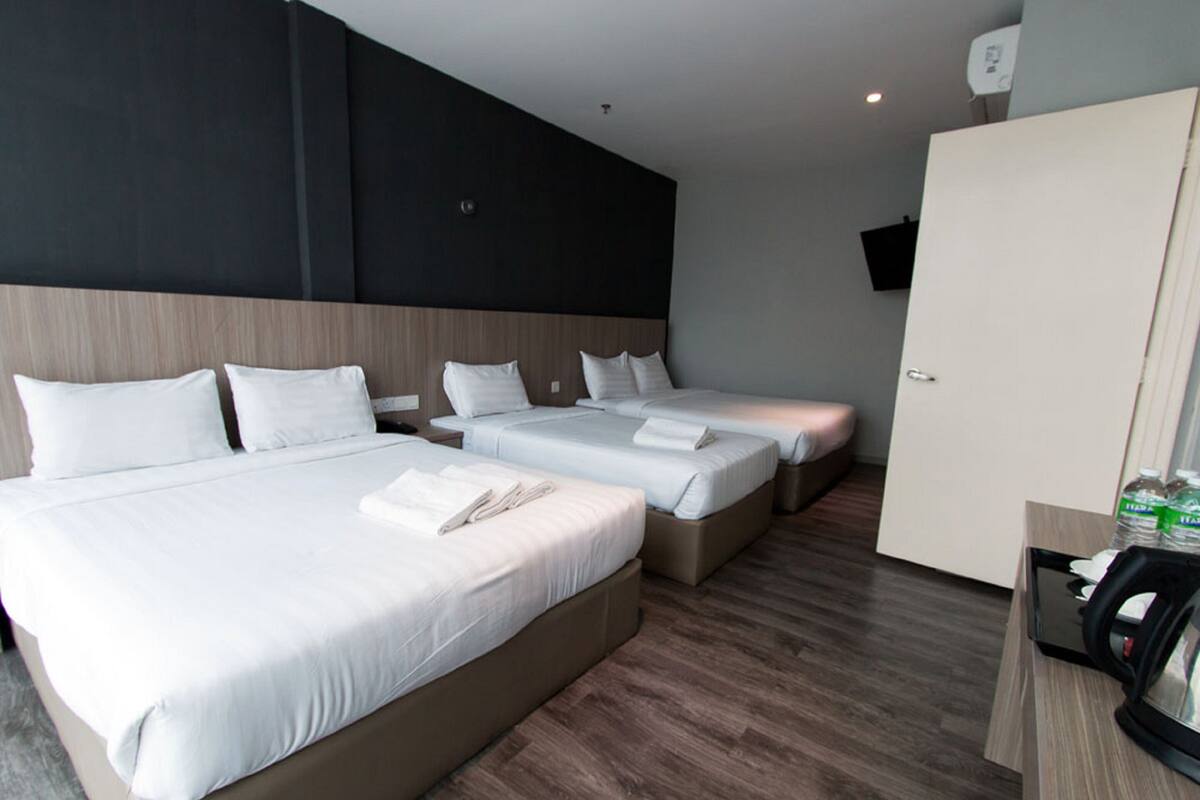 Een hotelkamer met een groot bed, twee kleinere bedden, een kledingkast, een bureau met een waterkoker, en een wandgemonteerde flatscreen-tv.
