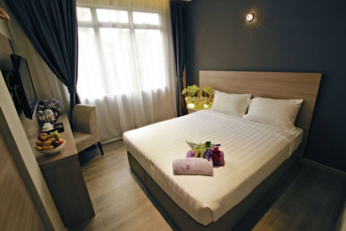 Een opgemaakt bed met een witte deken en kussens, een nachtkastje met een fruitschaal, een stoel en een vaas met bloemen.