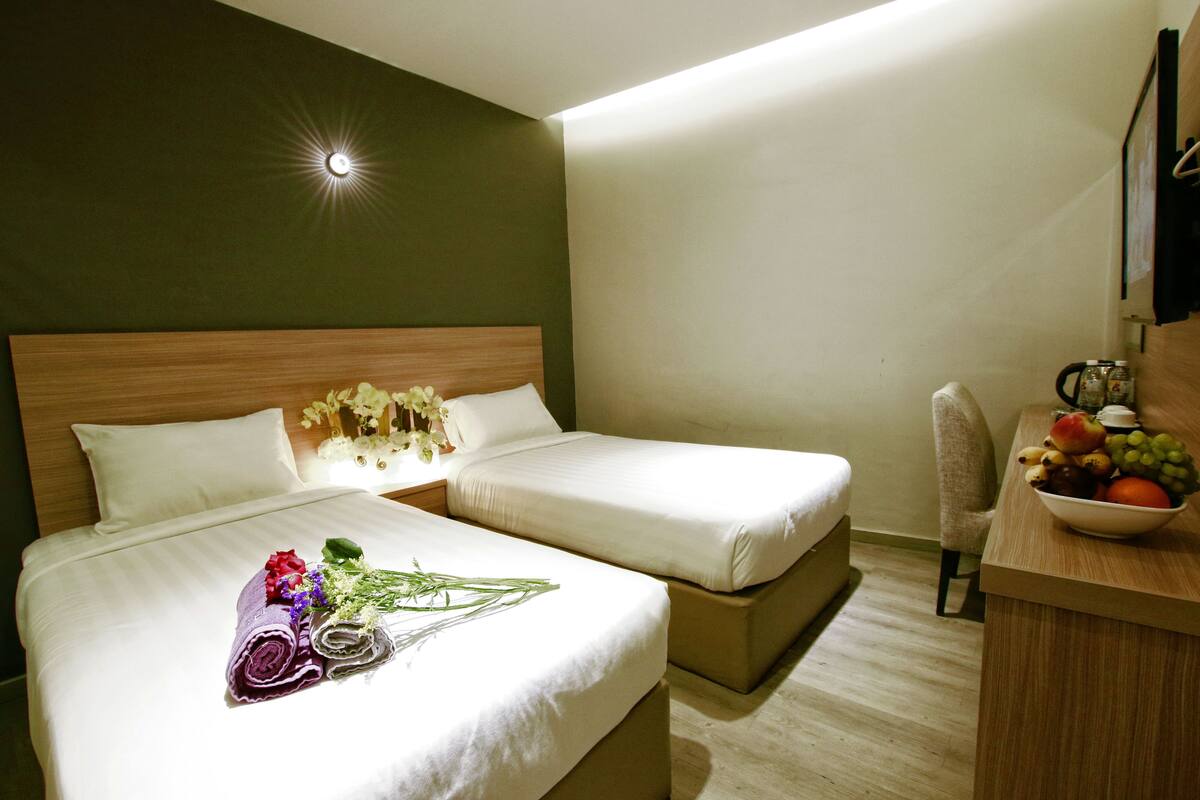 Een hotelkamer met twee bedden, een bureau met een schaal fruit en een aan de muur bevestigde televisie.