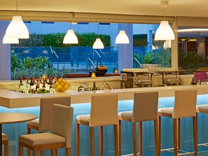 A bar area with multiple seating options, a bar counter with various bottles and glasses, and a view of greenery outside. - Novotel Rio de Janeiro Parque Olímpico (Rio de Janeiro)