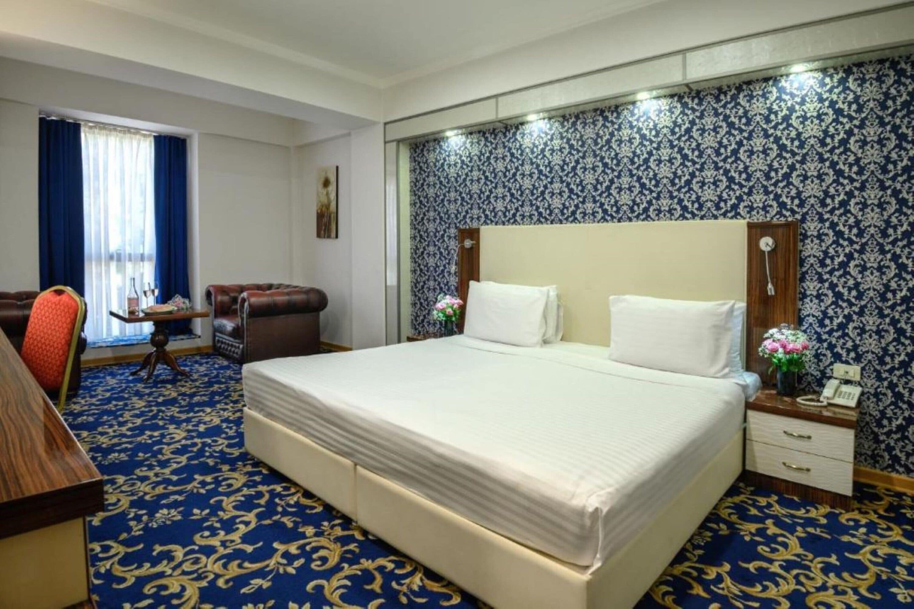 Une chambre d’hôtel avec un grand lit, un bureau avec une chaise, une petite table avec un vase et une moquette à motifs.