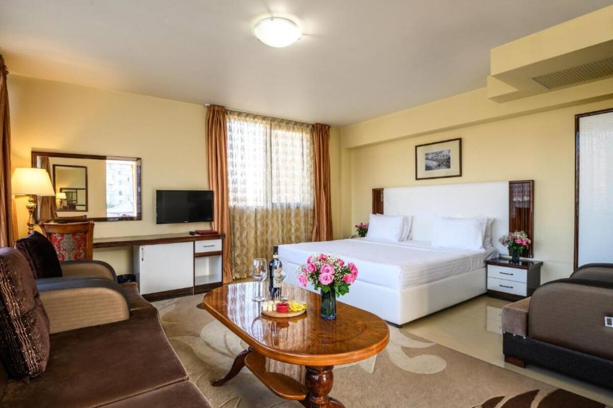 A hotel room with a bed, a desk, a TV, a sofa, and a coffee table with flowers.