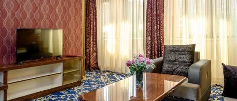A hotel room with a flat-screen TV, a wooden coffee table with a vase of flowers, a sofa, and a patterned carpet.