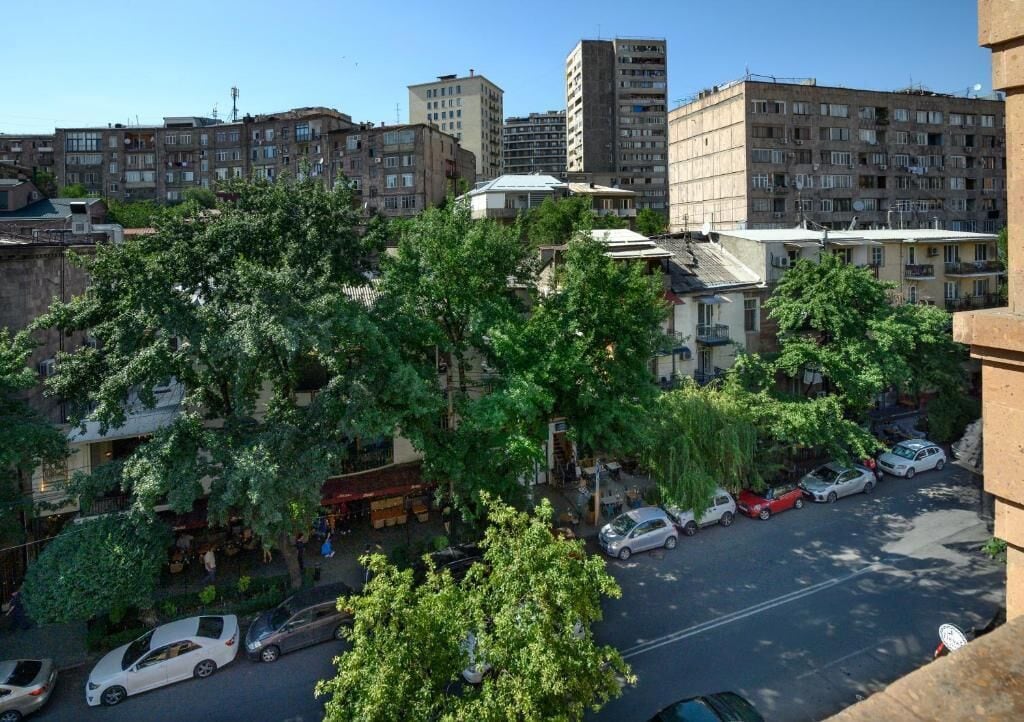 An urban street scene with multi-story buildings, parked cars, and trees.