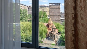 Blick aus einem Fenster mit Vorhängen. Im Vordergrund ein Gebäude mit einer Kuppel und ein kleineres Gebäude mit einem spitz zulaufenden Dach, umgeben von Grünflächen und weiteren Gebäuden im Hintergrund.