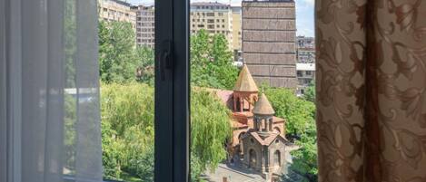Blick aus einem Fenster mit Vorhängen. Im Vordergrund ein Gebäude mit einer Kuppel und ein kleineres Gebäude mit einem spitz zulaufenden Dach, umgeben von Grünflächen und weiteren Gebäuden im Hintergrund.