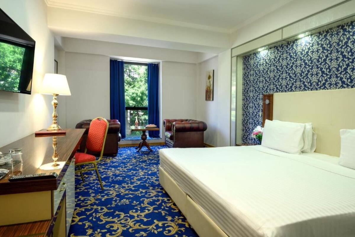 A hotel room with a large bed, a desk with a lamp, a chair, and a view of greenery outside.
