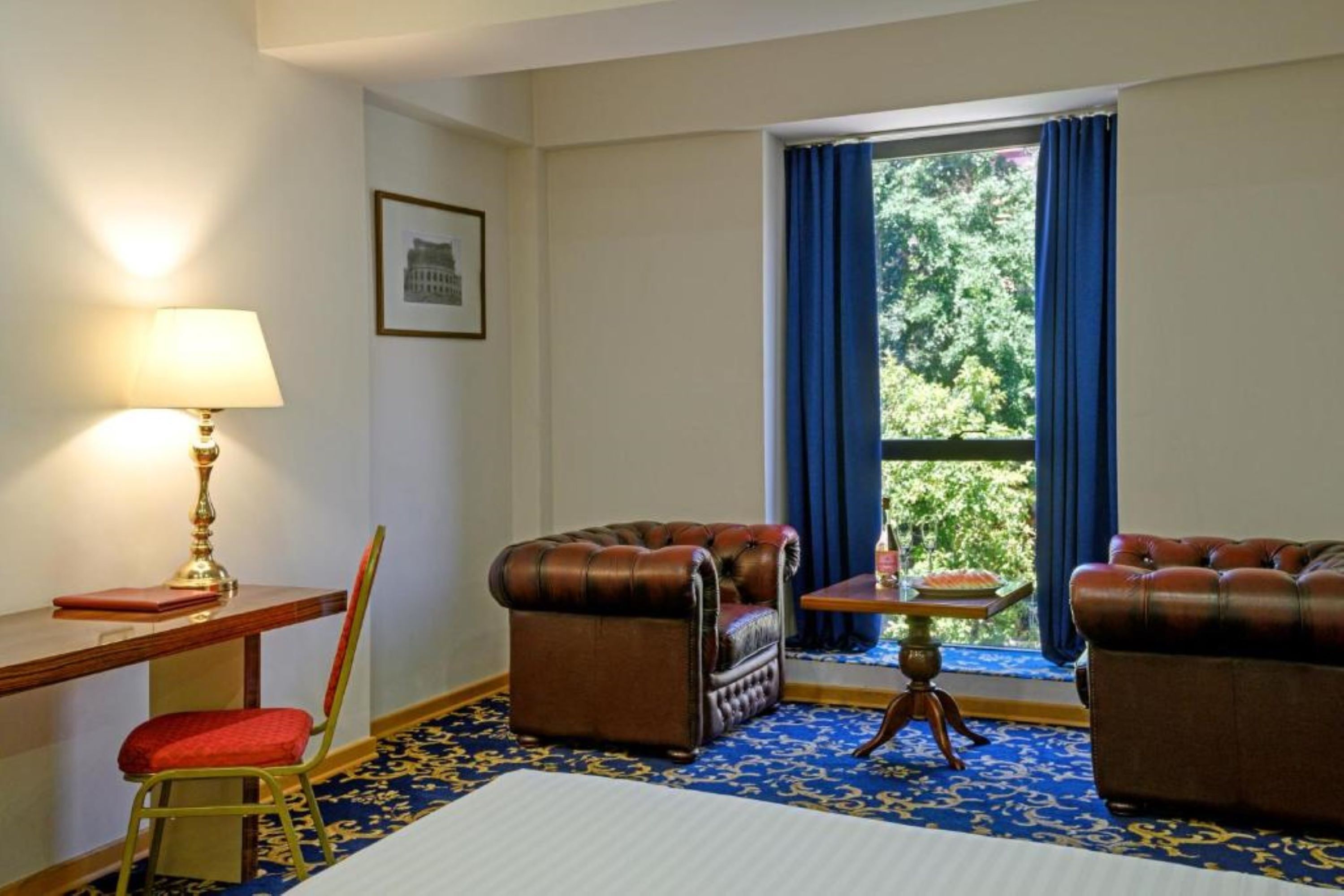 A room with two leather armchairs, a wooden desk with a lamp, a small table with a vase, and a window with blue curtains.