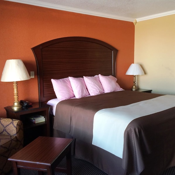 A hotel room with a wooden headboard, a bed with pink and white bedding, two bedside lamps, a small wooden table, and a patterned armchair.