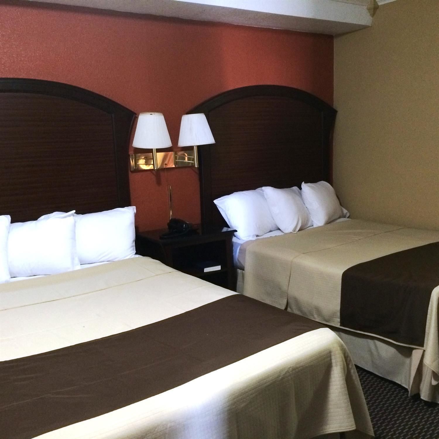 A hotel room with two beds, each with white linens and a brown headboard. There are two wall-mounted lamps on the wall between the beds.