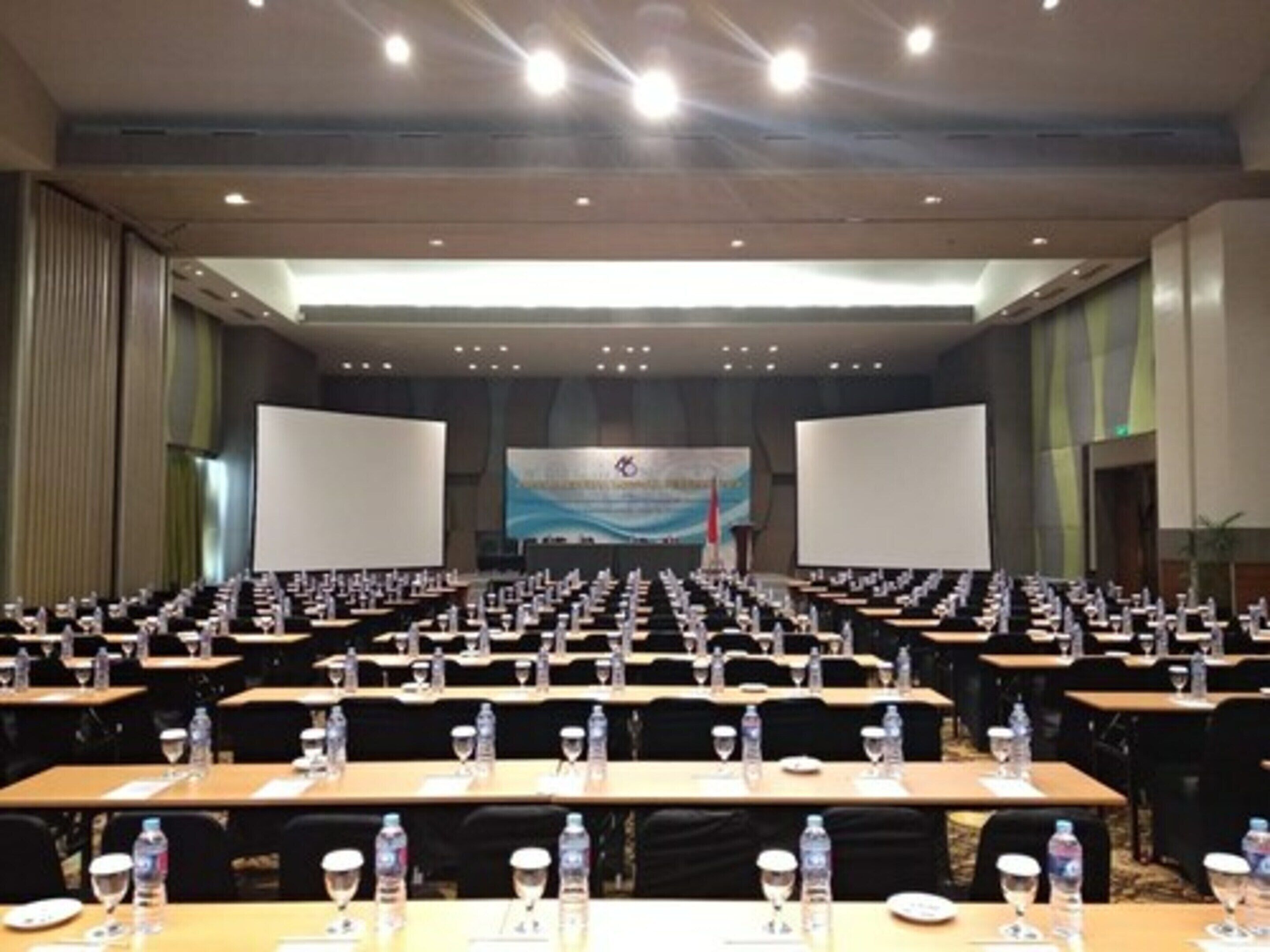 Un salón de conferencias con filas de mesas, cada una con botellas de agua y vasos, y una pantalla grande al frente.